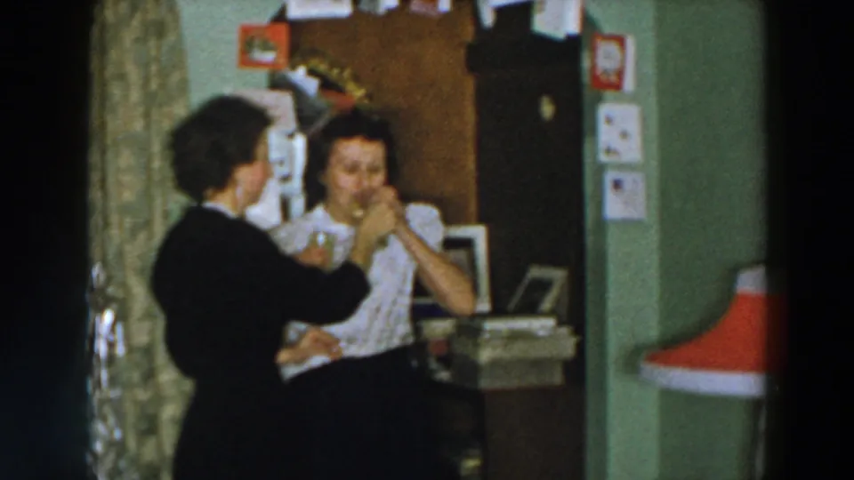 1957 Wheeling, Ohio frame showing two women interacting with a drink beside holiday tinsel, wall cards, and a red lampshade.