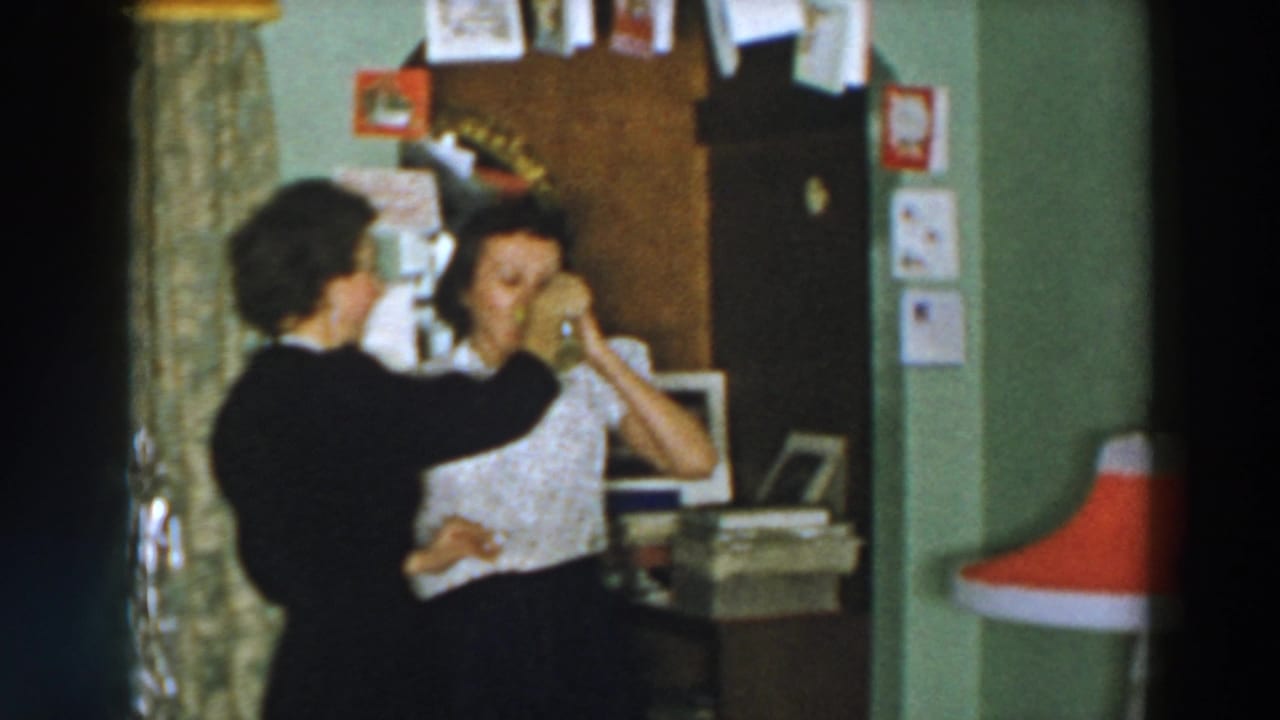 Two women indoors as one raises a patterned mug toward the other's face.