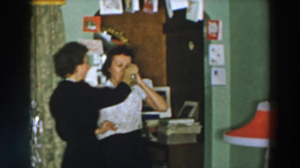 In a 1957 Wheeling Ohio home-movie frame, a woman in a dark dress offers a patterned mug to a woman in a white blouse indoors.