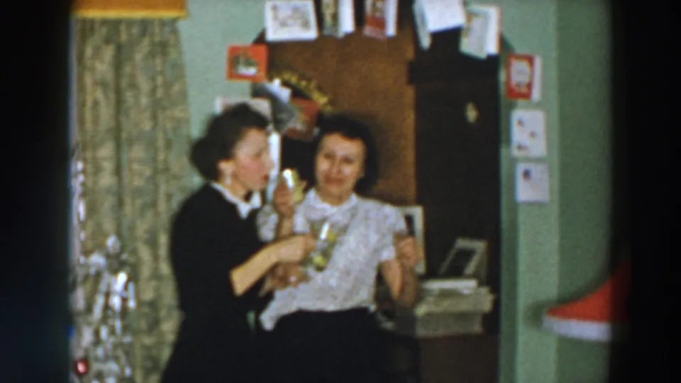A 1957 Wheeling Ohio home-movie still shows two women close together, laughing and holding mugs in a private interior.