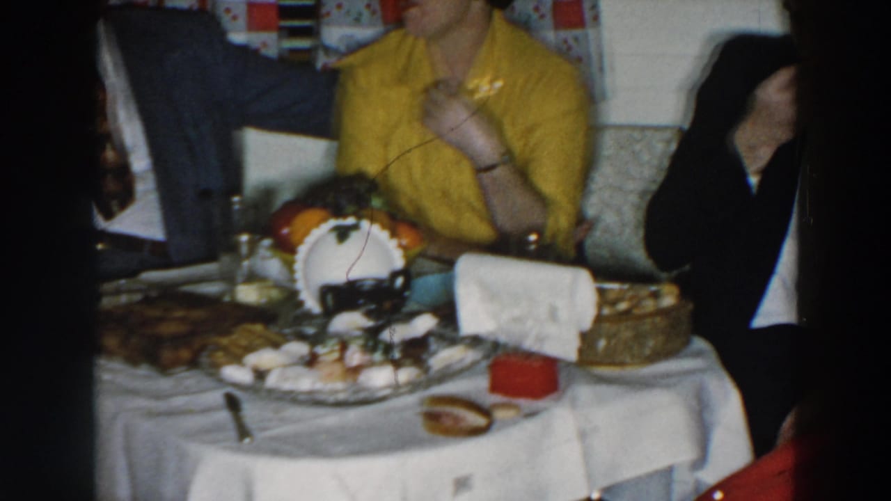 Adults seated at a dinner table with food, curtains, and a woman in a yellow blouse.