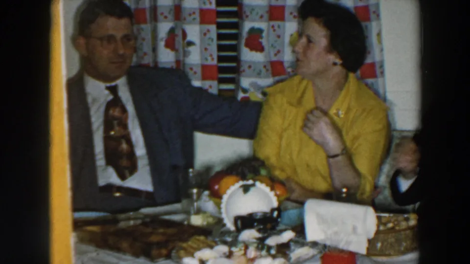 1957 Wheeling, Ohio dinner scene with a suited man and woman in yellow seated beside a food-covered table.