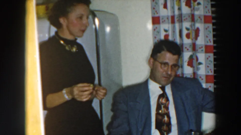 1957 Wheeling, Ohio interior frame with a standing woman near a refrigerator and a seated man in a suit.