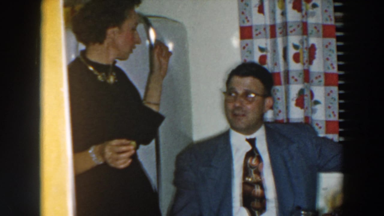 Woman standing beside a seated man in a grey suit at a kitchen table.