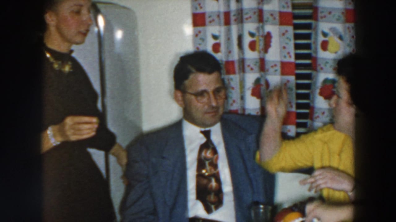 1957 Wheeling, Ohio kitchen scene with a woman in yellow gesturing toward a man in a grey suit as another woman stands near the refrigerator.