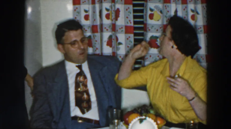 1957 Wheeling, Ohio frame showing a woman in a yellow blouse holding food near her mouth while a man in a grey suit watches at a kitchen table.