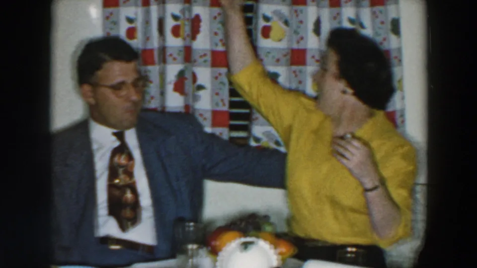 1957 Wheeling, Ohio home-movie frame with a woman in yellow raising her arm beside a man in a grey suit at a kitchen table with fruit and glasses.