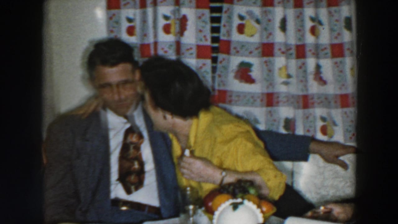 In a 1957 Wheeling, Ohio home-movie frame, a woman in a yellow blouse leans close to a man in a suit beside patterned curtains.