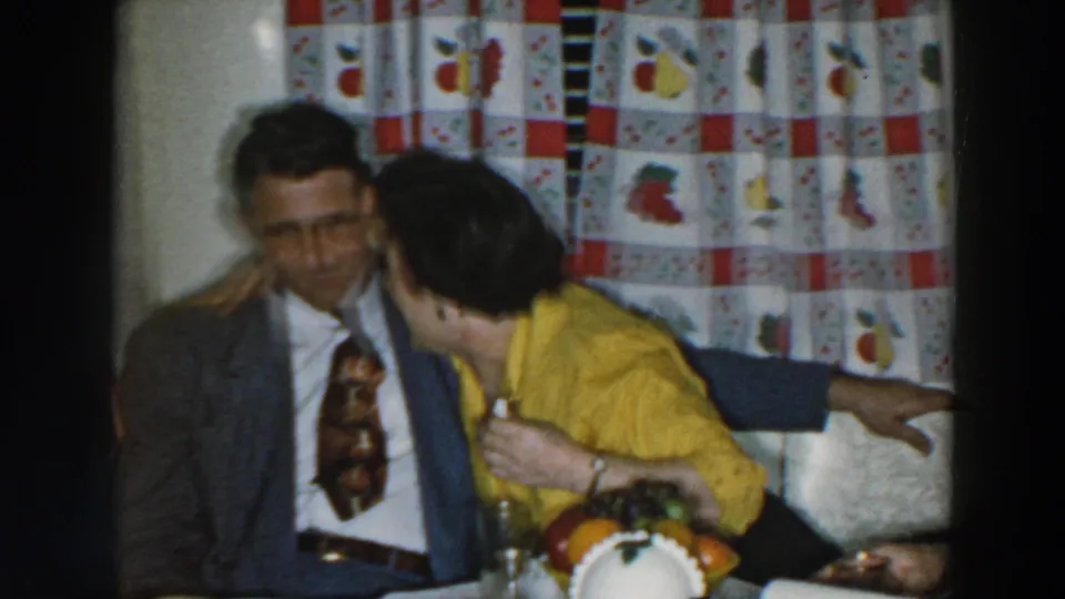 In a 1957 Wheeling, Ohio home-movie frame, a woman in a yellow blouse leans close to a man in a suit beside patterned curtains.