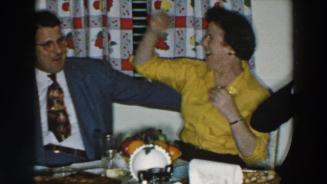 Woman in a yellow blouse raises her arm while seated beside a man in a blue suit.