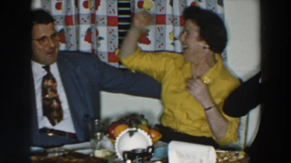 The woman in yellow gestures playfully beside a seated man in a blue suit at a table in the Wheeling home-movie scene.