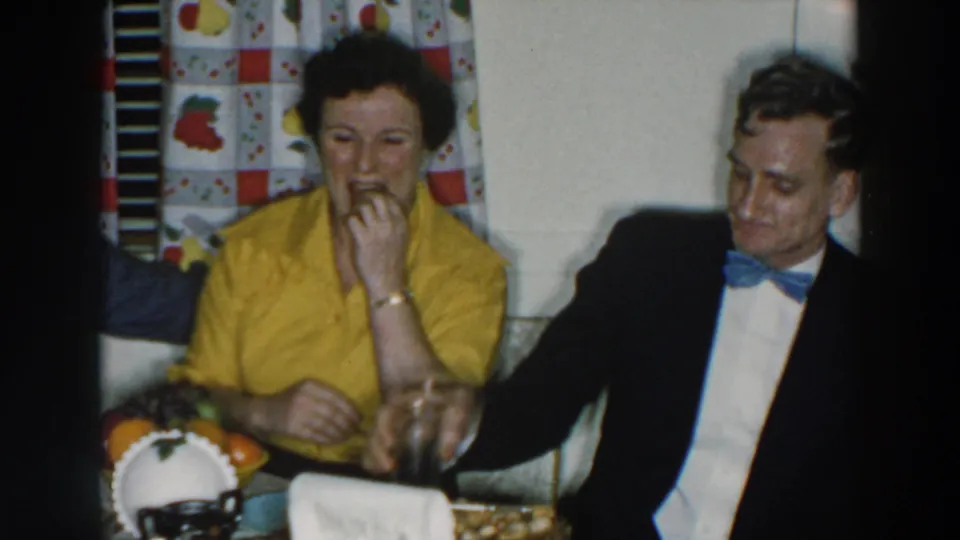 At the indoor gathering, the woman in yellow eats while a man in a dark jacket and blue bow tie sits beside her.