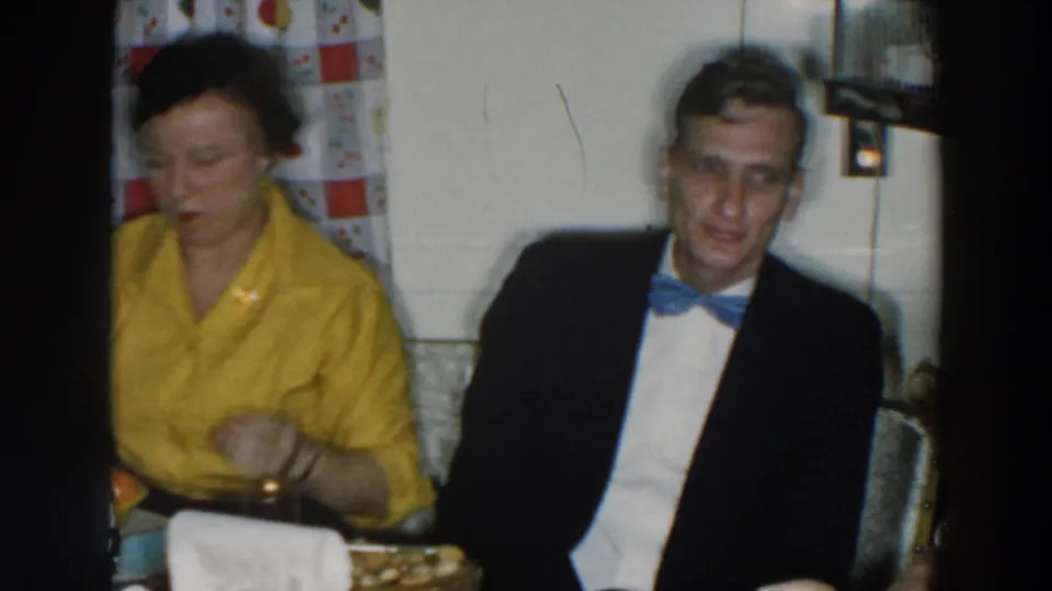 The final frame shows the woman in yellow and a bow-tied man seated at the table, with curtains and dishes behind the home-movie action.