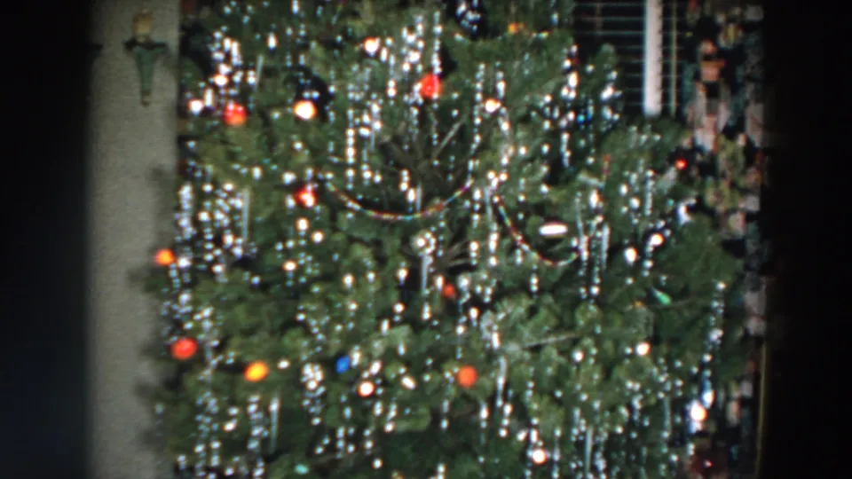 The 1957 home-movie frame centers on a heavily decorated Christmas tree with reflective tinsel, colored ornaments, and soft interior lighting.