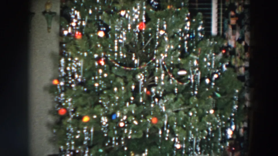 A close 8mm view of the Wheeling Christmas tree shows dense greenery, dangling silver tinsel, red and orange ornaments, and dim room surroundings.