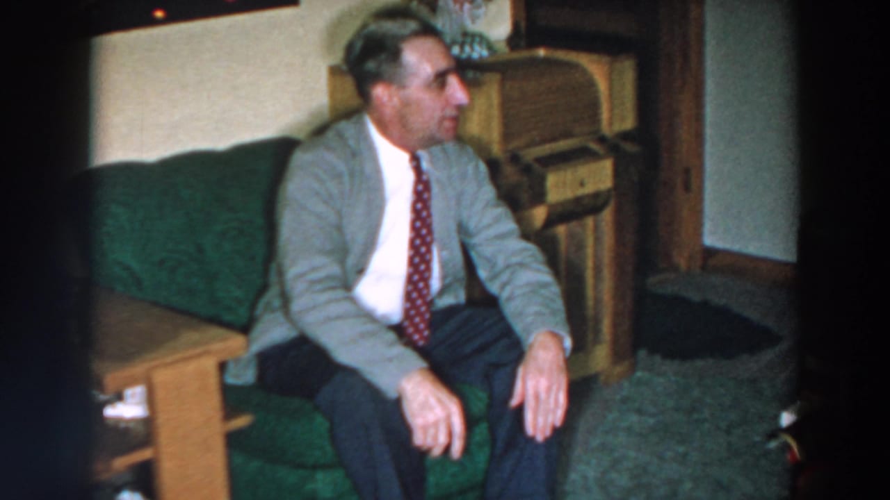 In 1957 Wheeling, Ohio metadata, a man in a gray blazer and red tie sits on a green couch beside wood furniture.