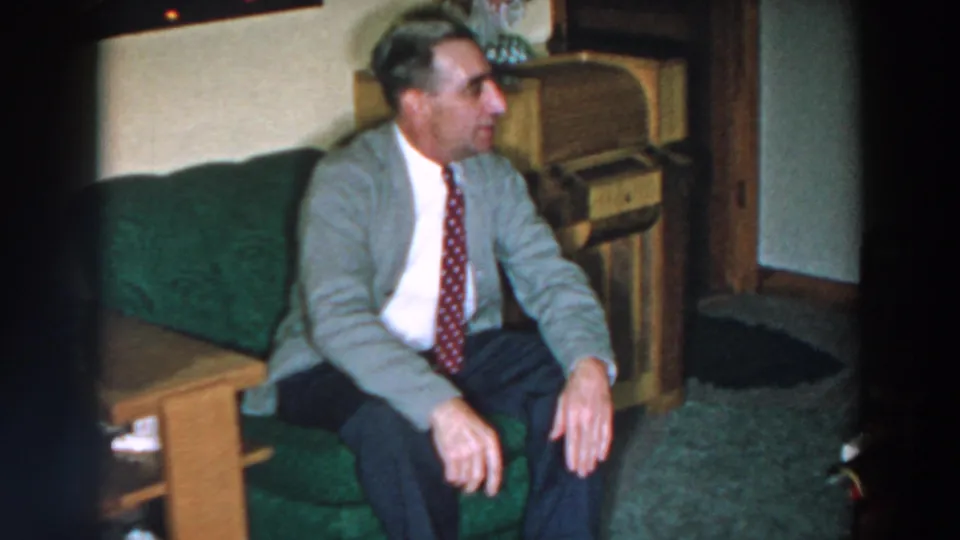 In 1957 Wheeling, Ohio metadata, a man in a gray blazer and red tie sits on a green couch beside wood furniture.