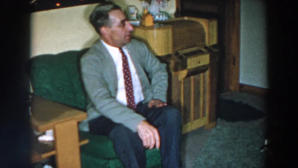 The seated man leans back on the green couch in the Wheeling, Ohio 1957 home-movie scene, with wood furniture behind him.