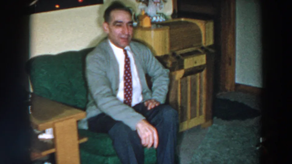 The man turns more toward the camera in the 1957 home interior, seated on a green couch with a cabinet and carpet visible.