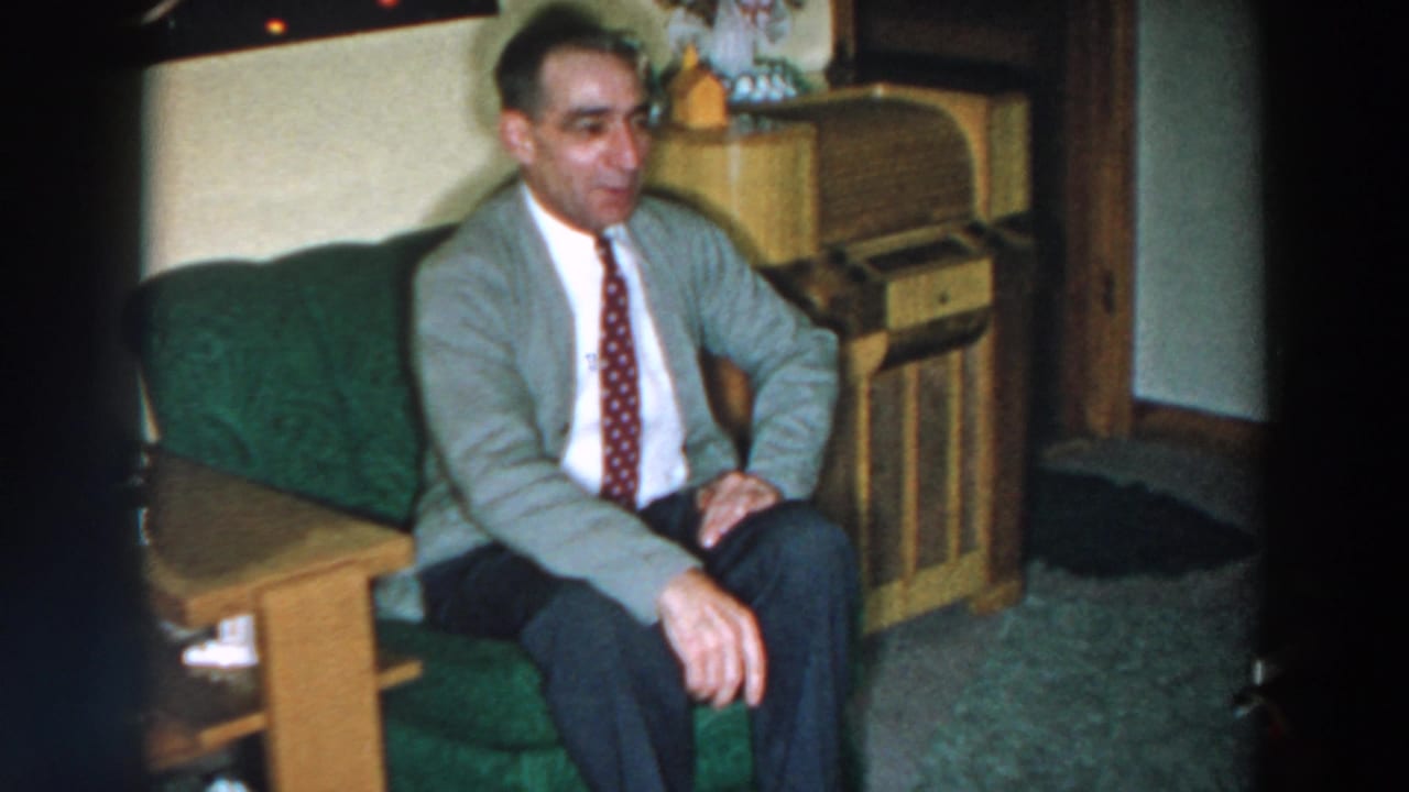 In 1957 home-movie footage from Wheeling, Ohio, a man in a gray cardigan and patterned tie sits on a green couch beside a wooden cabinet.