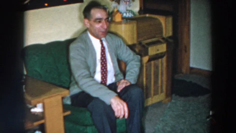 In 1957 home-movie footage from Wheeling, Ohio, a man in a gray cardigan and patterned tie sits on a green couch beside a wooden cabinet.