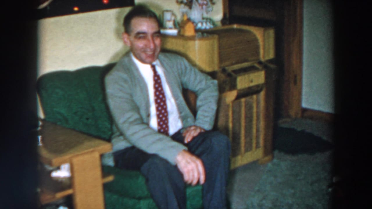 Smiling man sits on a green couch beside a wooden cabinet.