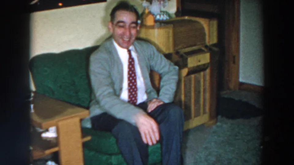 A seated man in a gray cardigan lifts his hand in a friendly gesture in a 1957 home-movie living-room scene.
