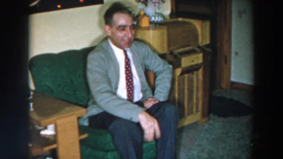The man remains seated on the green couch, smiling and gesturing toward the camera in the 1957 Wheeling, Ohio home-movie clip.