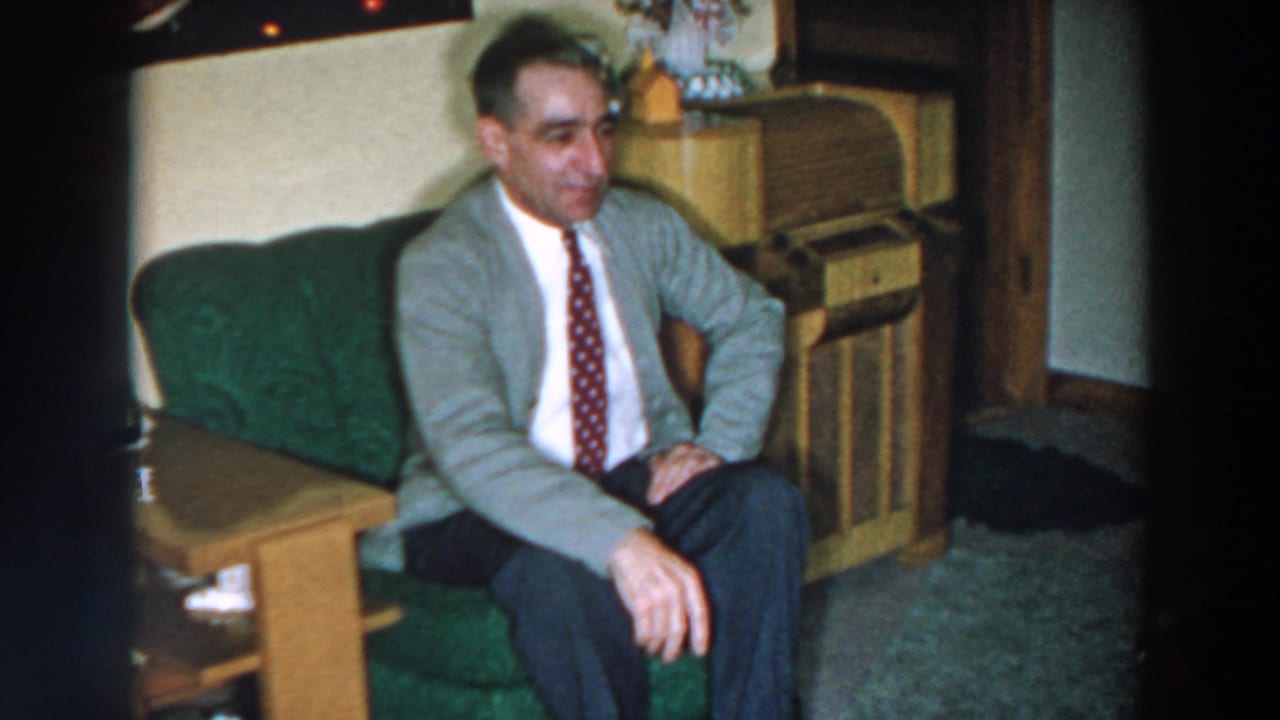 Man seated on a green sofa in a living room beside a wooden table and console cabinet.