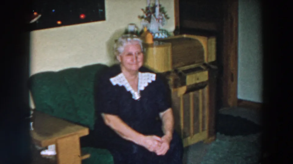 In supported 1957 Wheeling, Ohio footage, a woman in a dark dress with a white lace collar sits on a green sofa in a living room.