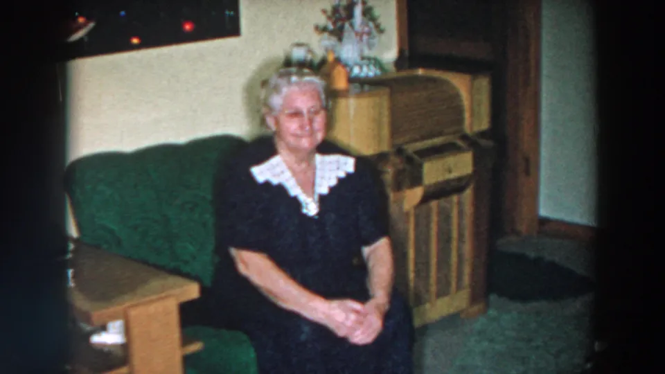 The 1957 Wheeling, Ohio home-movie frame shows a woman seated on a green sofa near a wooden console cabinet in a private living room.