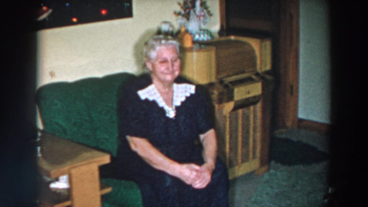 In a 1957 Wheeling, Ohio home-movie frame, an elderly woman smiles from a green sofa beside a wooden cabinet.