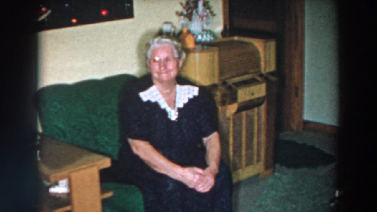 Elderly woman smiling at the camera with hands clasped on a green sofa.