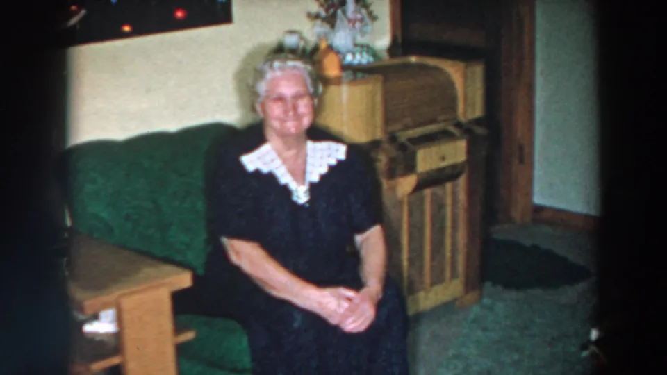 A 1957 home-movie frame shows the seated woman on a green sofa, with a wooden cabinet and holiday-like decor behind her.