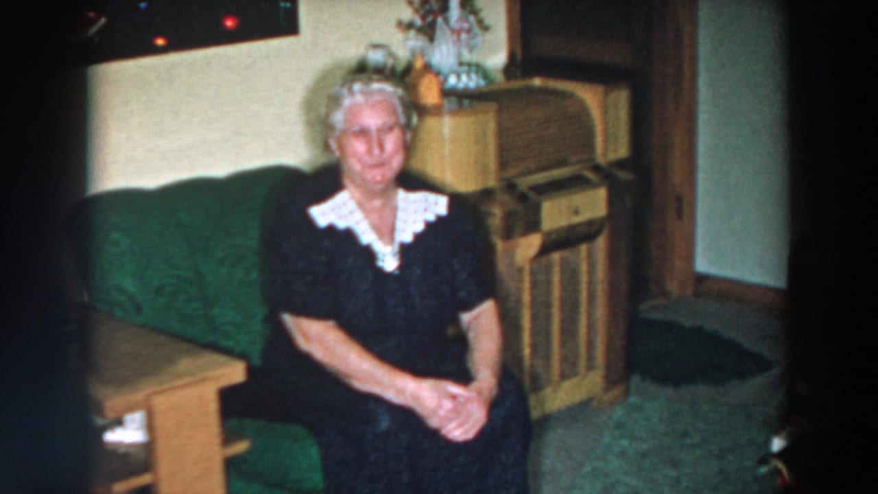 Elderly woman seated on a green couch beside a wooden table and console radio.