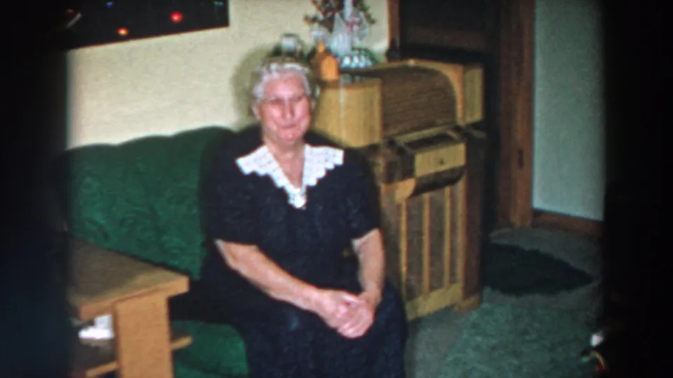 Elderly woman in a dark dress with a white collar sits on a green couch in a 1957 Wheeling, Ohio living room.