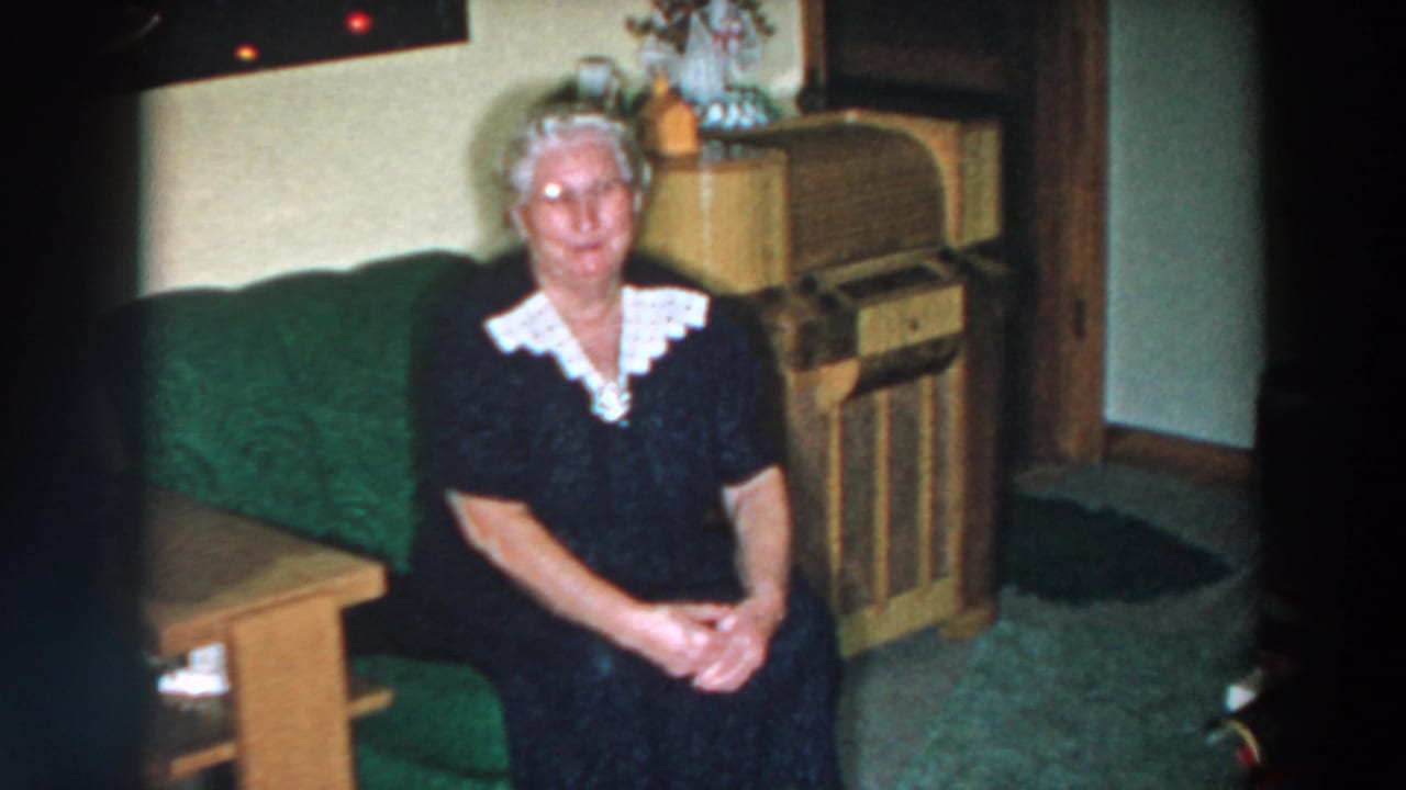 Woman seated with folded hands in a living room with a radio cabinet behind her.