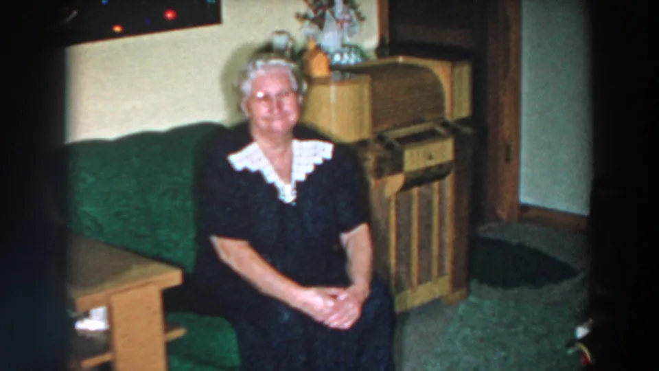 Elderly woman sits upright on a green couch in a 1957 Wheeling, Ohio living room with wood furniture and soft 8mm color.
