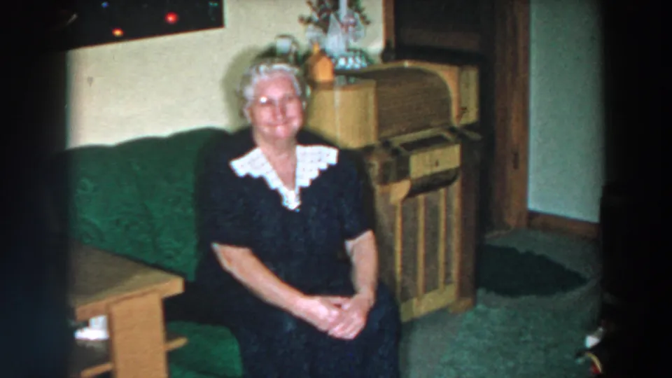 Elderly woman smiles at the camera from a green couch in a 1957 Wheeling, Ohio domestic interior.