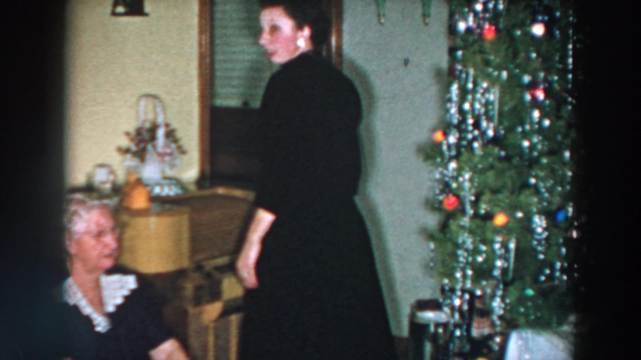Adults in a living room near a decorated Christmas tree, with an older woman seated in the foreground.