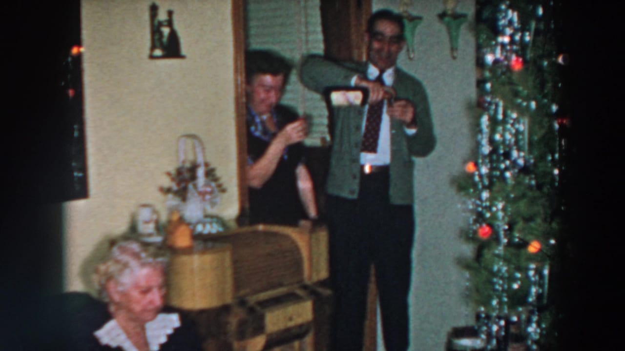 A man in a green cardigan pours a drink while adults stand near a Christmas tree.