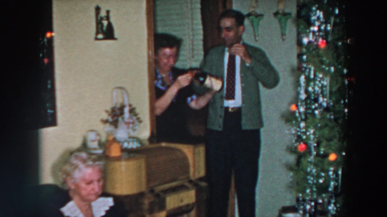Adults stand with drinks near a decorated Christmas tree while an older woman sits in the foreground.