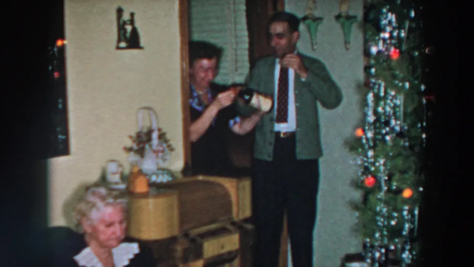 1957 Wheeling, Ohio home-movie frame showing adults with drinks beside a tinsel Christmas tree and an older seated woman.