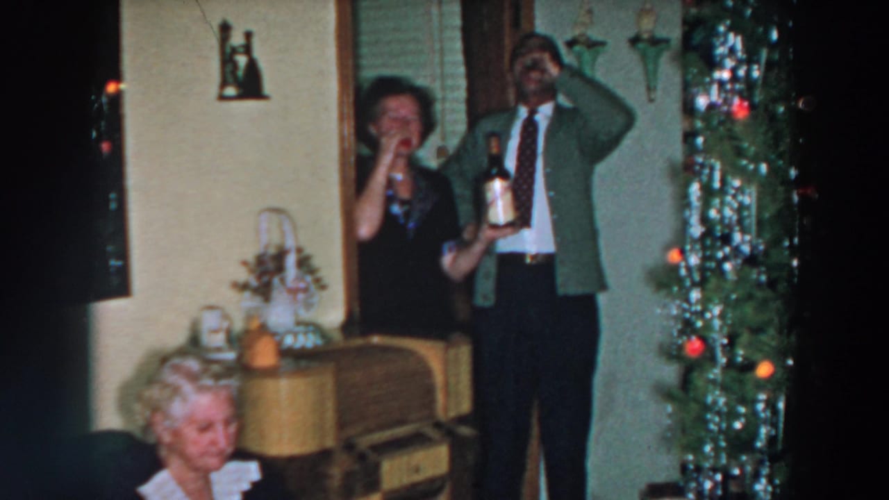 A man raises a glass as a woman holds a bottle near a decorated Christmas tree.
