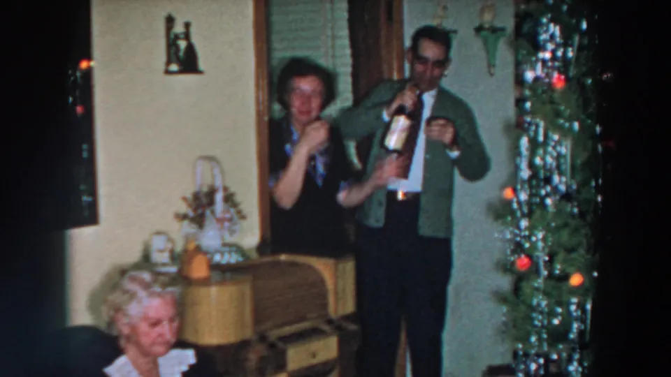 Home-movie frame from 1957 Wheeling, Ohio showing a smiling woman with a glass and a man pouring or handling alcohol.