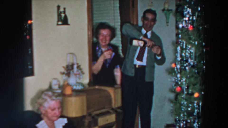 1957 Wheeling, Ohio home-movie frame of a man pouring alcohol as a woman smiles near a lit, tinsel-covered Christmas tree.