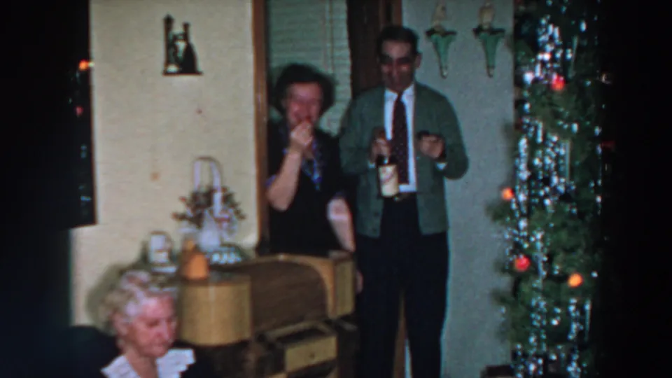 A woman raises a drink as a man stands with a bottle and glass in a Wheeling, Ohio holiday home scene from 1957.
