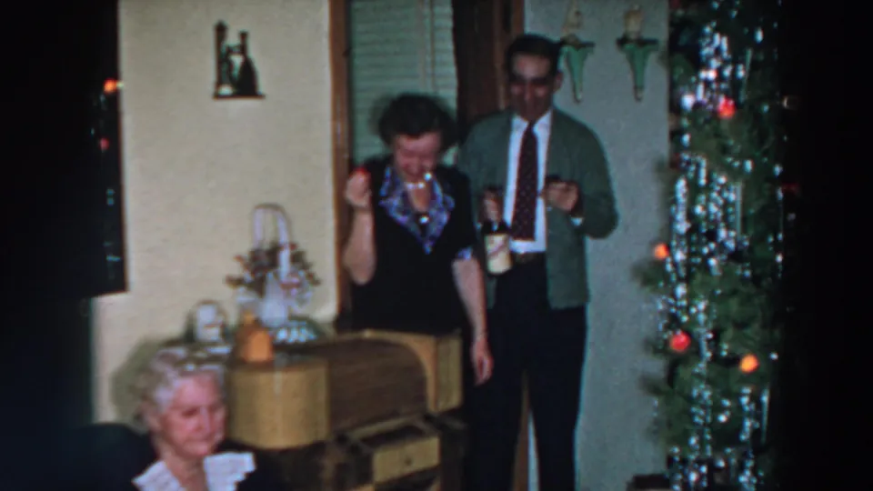 The woman lowers her gaze while holding a glass, with a man and decorated Christmas tree behind her in the 1957 home-movie scene.
