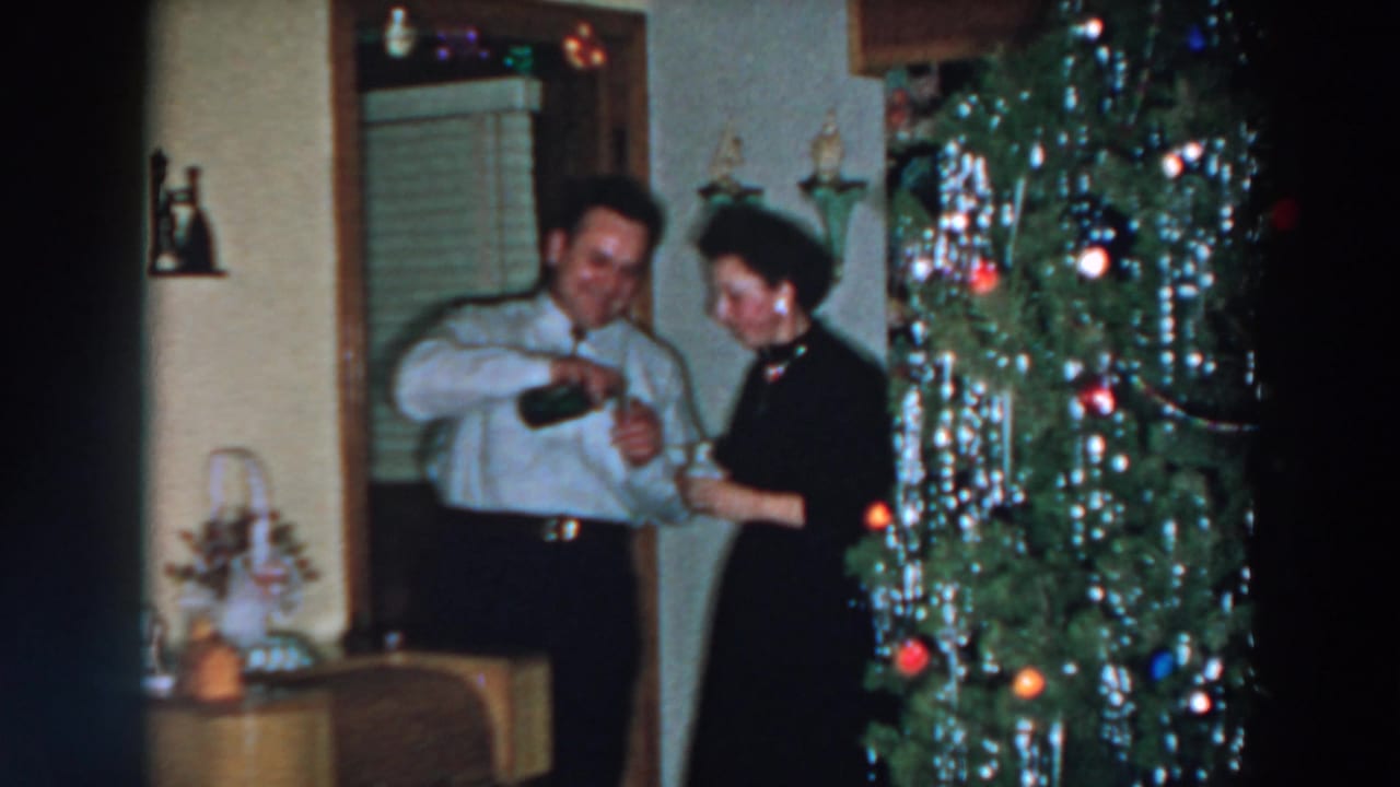 Man and woman stand beside a decorated Christmas tree while handling a bottle and glass.