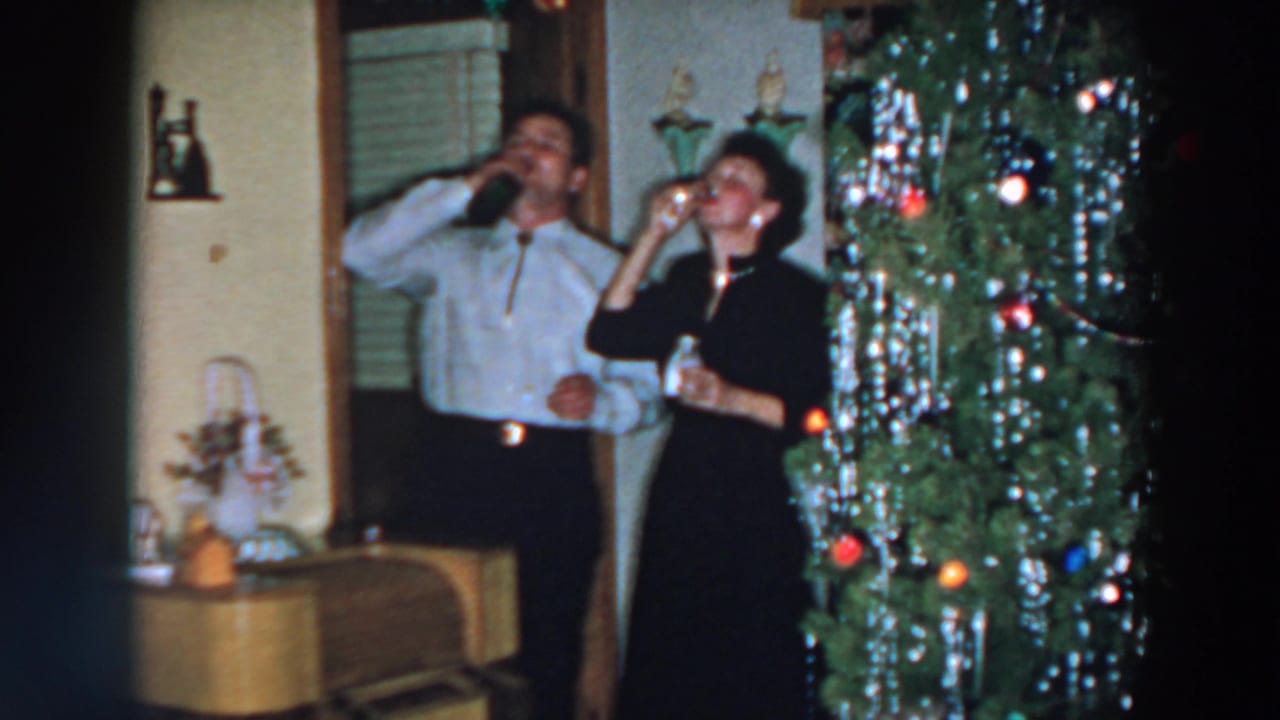 Man and woman drink beside a decorated Christmas tree in a private home.